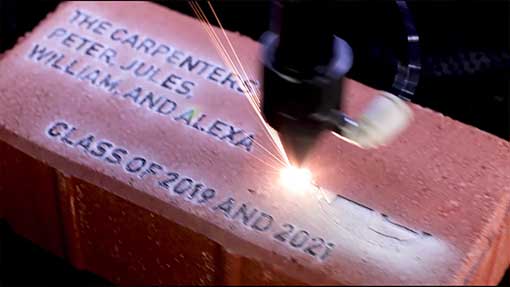 Laser engraving machine in action, etching the names 'THE CARPENTERS PETER JULES, WILLIAM, AND ALEXA CLASS OF 2019 AND 2021' onto a brick