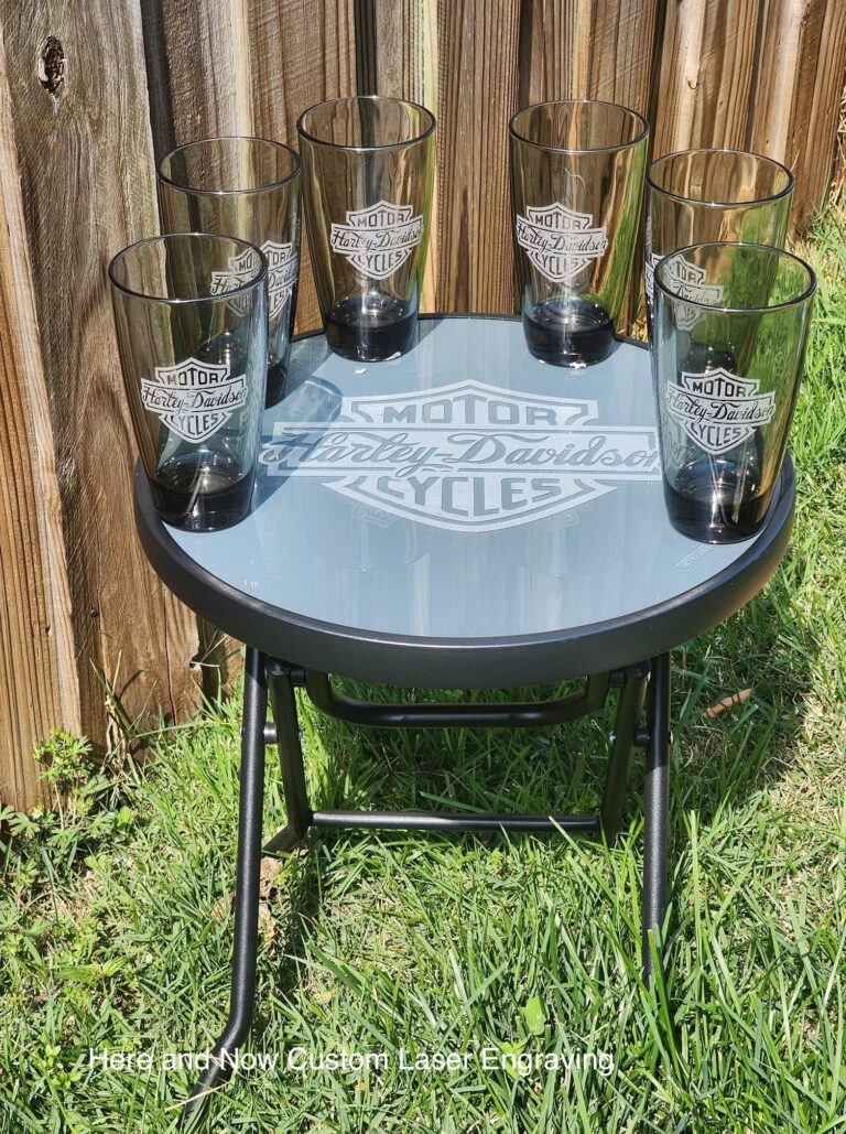 A custom-engraved glass-top table and matching drinkware set featuring the Harley-Davidson logo, created by Here and Now Custom Laser Engraving, displayed outdoors against a wooden fence.