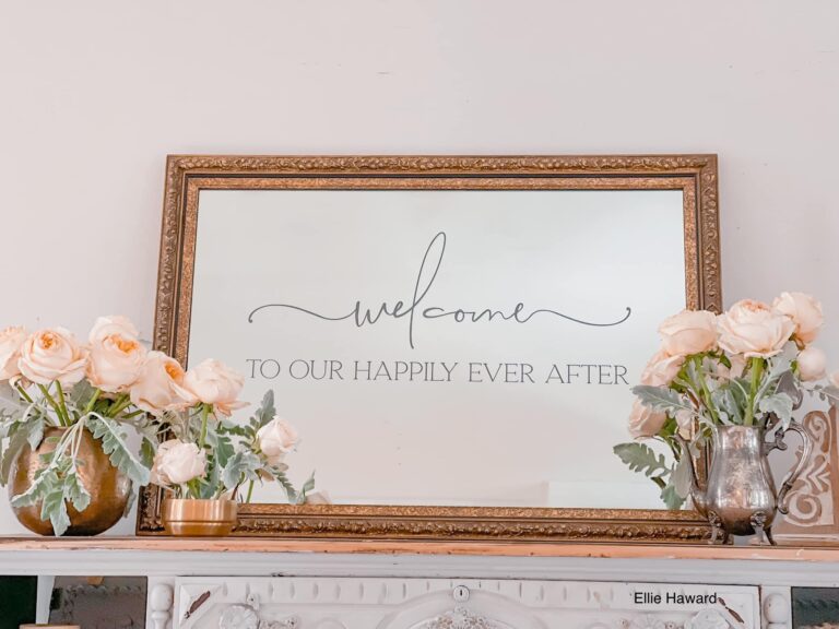 A beautifully engraved mirror with a gold frame displaying the message "Welcome, We Are So Happy You Are Here," styled in an elegant interior setting with vintage furniture and soft floral arrangements.