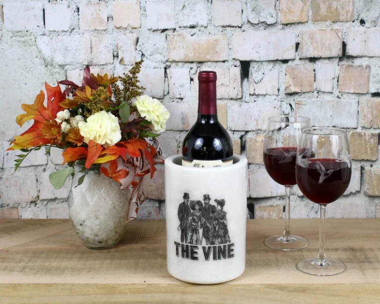 A custom-engraved wine chiller with the text "THE VINE" and a vintage illustration, accompanied by two engraved wine glasses filled with red wine, set against a rustic brick wall with a floral arrangement.